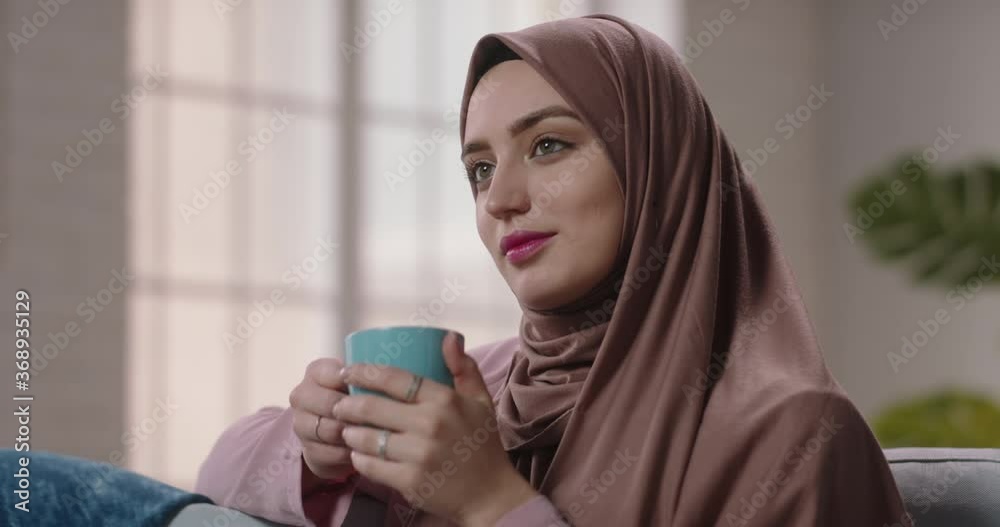 Beautiful middle eastern model in traditional hijab head scarf sitting at home, drinking tea or coffee and positively smiling - beauty, modern muslim concept 4k footage Beautiful middle eastern model in traditional hijab head scarf sitting at home, drinking tea or coffee and positively smiling - beauty, modern muslim concept 4k footage