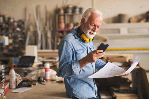 Likeable older carpenter is looking at the cellphone and smiling photo