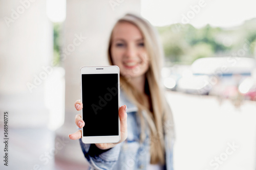 Close up portrait of female happy smiling student showing a screen of a mobile phone outdoor on the background of a university building