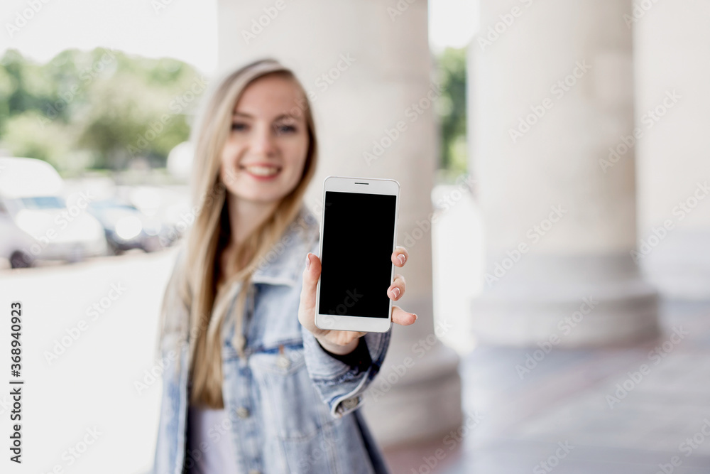 Happy blonde girl showing mobile phone and smiling on the background of a university, mockup for design, copy space Happy blonde girl showing mobile phone and smiling on the background of a university, mockup for design, copy space