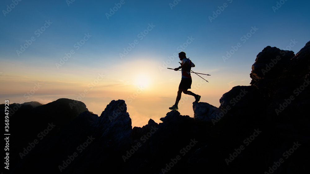 Sunset silhouette of skyrunner man descending from alpine ridge with poles