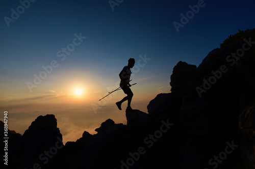 Sunset silhouette of skyrunner man climbing alpine ridge with poles