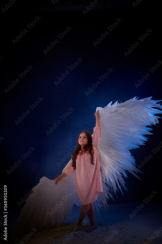 Naklejka premium Small young model girl with white wings posing and dancing in dark black studo during photoshoot with flour or dust and colour light. Actress in performance about struggle between good and evil.
