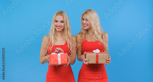 The twin girls in red show off gifts in boxes to each other smiling and then to the camera. The presentation of gift wrappings for the New Year holiday.