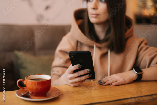 Close-up portrait of young attractive woman typing message in phone at cozy coffee shop. Pretty girl in casual clothes thinks while typing on the phone at cafe. Concept of leisure activity.