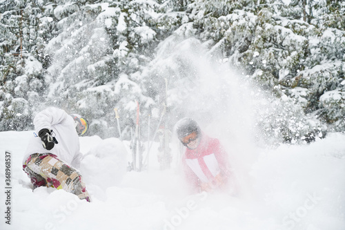 Young couple skiers in ski suits and helmets throwing fresh powder snow in the air and smiling. Man and woman in ski goggles having good time at ski resort with beautiful snowy trees on background.