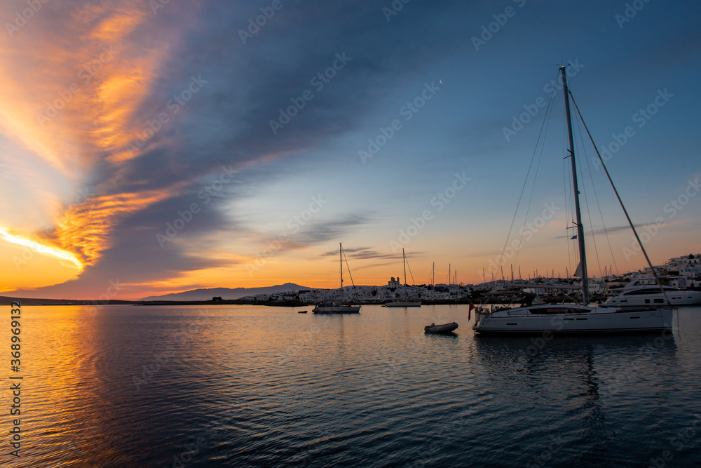 Anchored sailboats with magnificent sunrise with saturated orange and purple bank of clouds in the sky and moonset in Naoussa Village on Paros Island, Greece.