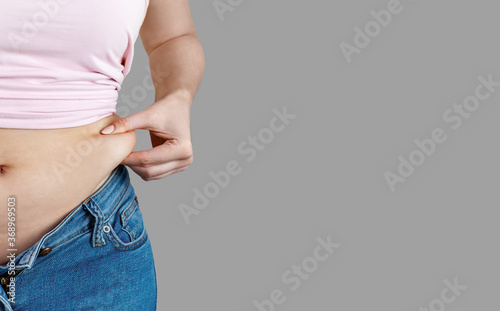 Woman in unbuttoned jeans holds squeezes excess belly fat closeup. The concept of overweight, weight loss, diet, unhealthy lifestyle, obesity, junk food on gray background, banner, copy space. © KseniyA