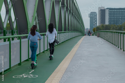 Photography Back view of two young women on their rental electric scooters on a bridge in Bratislava on their daily commute in cloudy weather