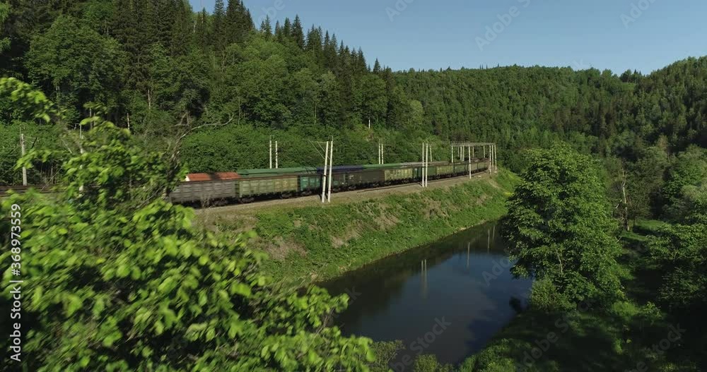 Two Freight trains with cargo and coal carriages an electric locomotive by two-sided Siberian railways near river in the forest mountains / Aerial drone mid view at summer sunset Two Freight trains with cargo and coal carriages an electric locomotive by two-sided Siberian railways near river in the forest mountains / Aerial drone mid view at summer sunset