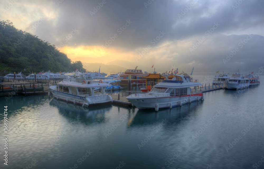 Tourist boats moored to the docks during partial solar eclipse at Shuishe Pier of Sun-Moon Lake in Nantou, Taiwan, with golden sun light shining through moody cloudy sky reflected on peaceful water