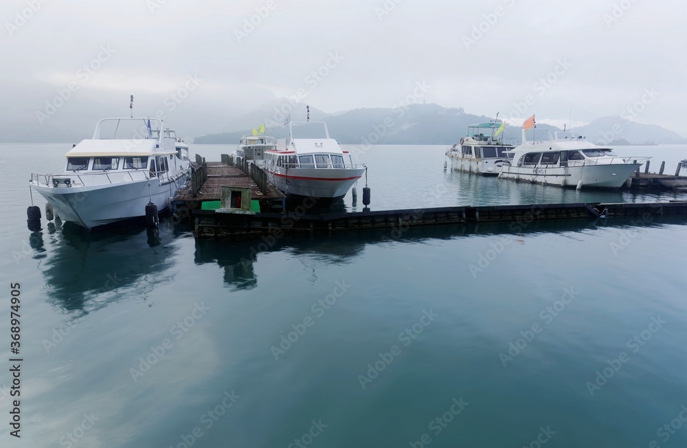 Tourist boats parking on peaceful water and moored to the floating docks of Shuishe Pier at Sun-Moon Lake on a foggy morning in Nantou, Taiwan, with mountains veiled in the fog under moody cloudy sky