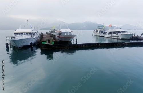 Tourist boats parking on peaceful water and moored to the floating docks of Shuishe Pier at Sun-Moon Lake on a foggy morning in Nantou, Taiwan, with mountains veiled in the fog under moody cloudy sky © AaronPlayStation