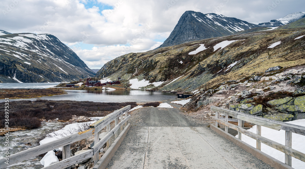 View overlooking Rondvassbu tourist cottages in Rondane national park. Wooden bridge in the foreground, gravel road and snow capped mountain peaks.