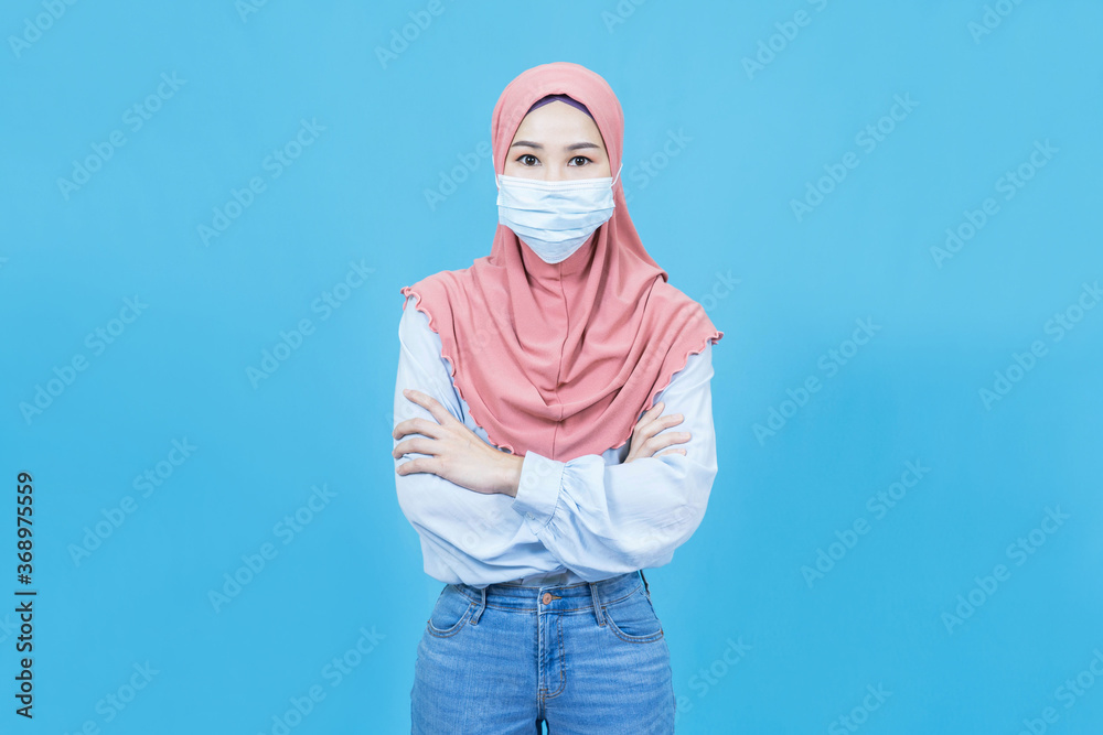 Portrait of young beautiful asian muslim woman in traditional dress wearing medical face mask folded arms influenza viral disease prevention stay home isolated over blue color background Portrait of young beautiful asian muslim woman in traditional dress wearing medical face mask folded arms influenza viral disease prevention stay home isolated over blue color background