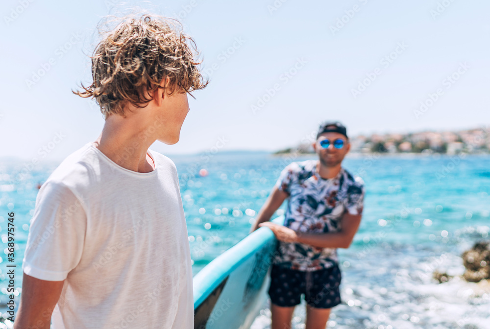 Fototapeta premium Blonde teenage son gazing to smiling father surfer carrying the surfing board on the bright sunny day noon. Active family summer vacation time near the ocean concept image.