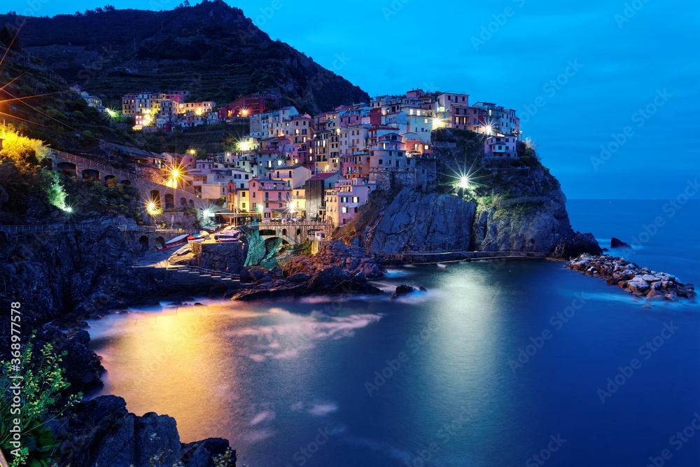 Night scenery of Manarola on vertical cliffs by the rocky coast with beautiful lights reflected on sea water, an amazing village in Cinque Terre National Park, Liguria, Italy, Europe (Long Exposure)