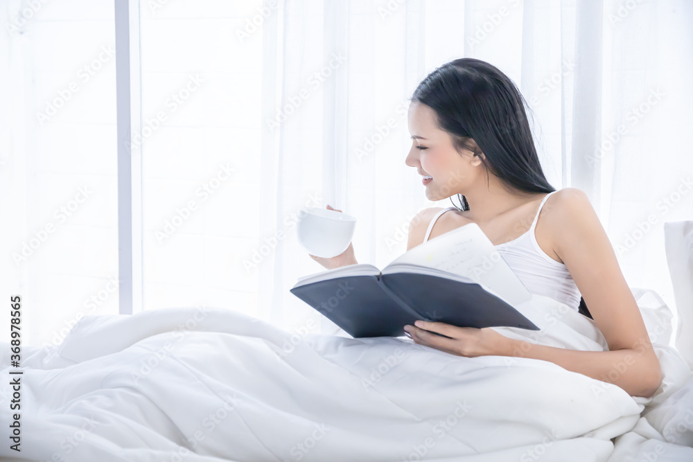 A Asian sexy girl sitting on white bed in bedroom and drinking a cup of hot coffee with reading a book, happy vacation morning