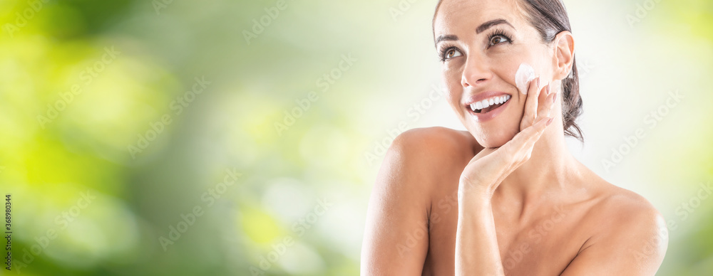 Beautiful young woman applying cream to her face. Skincare spa wellnes and cosmetics concept. Blurred green panoramic banner - obrazy, fototapety, plakaty 