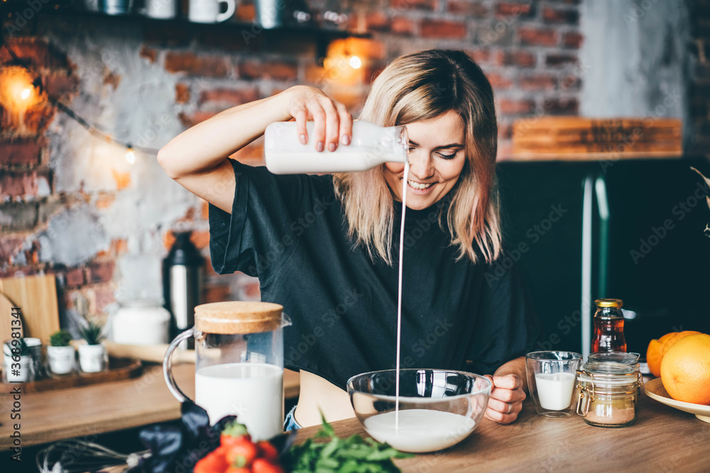 Young woman with fair hair pours milk from glass bottle into transparent bowl on wooden table with fresh fruits against orange brick wall at home. Young woman with fair hair pours milk from glass bottle into transparent bowl on wooden table with fresh fruits against orange brick wall at home.