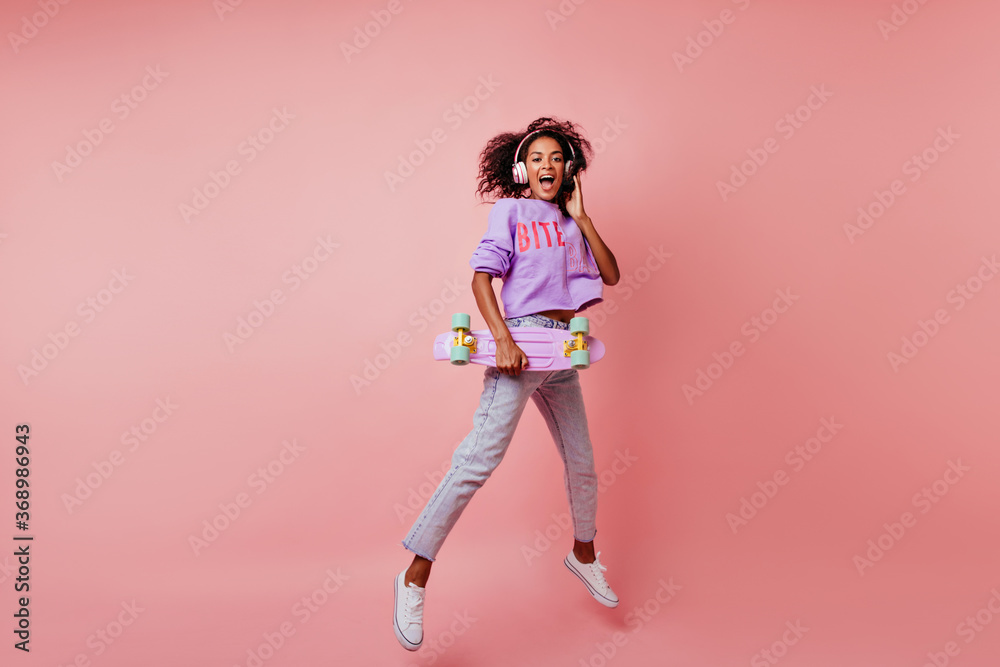 Full-length shot of stunning black woman in stylish jeans jumping on pink background. Attractive curly african girl with skateboard expressing positive emotions.