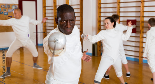 African American sportsman at fencing workout, training attack movements with rapier.