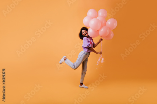 Graceful african female model in casual clothes fooling around on yellow background. Indoor photo of emotional birthday girl dancing with party balloons.