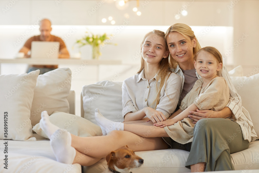 Warm-toned portrait of happy mother embracing two daughters while posing together sitting on couch in cozy home interior with father in background, copy space Warm-toned portrait of happy mother embracing two daughters while posing together sitting on couch in cozy home interior with father in background, copy space