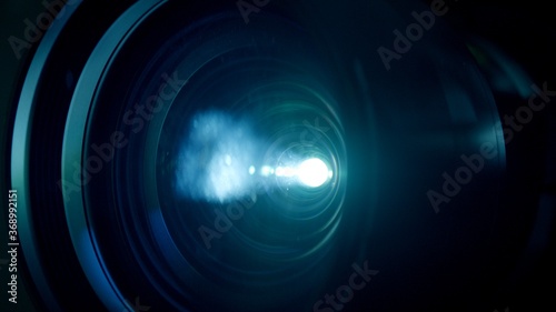 Macro closeup locked shot with selective focus of activated film projector. Illuminated lens and white light flare in dark room.