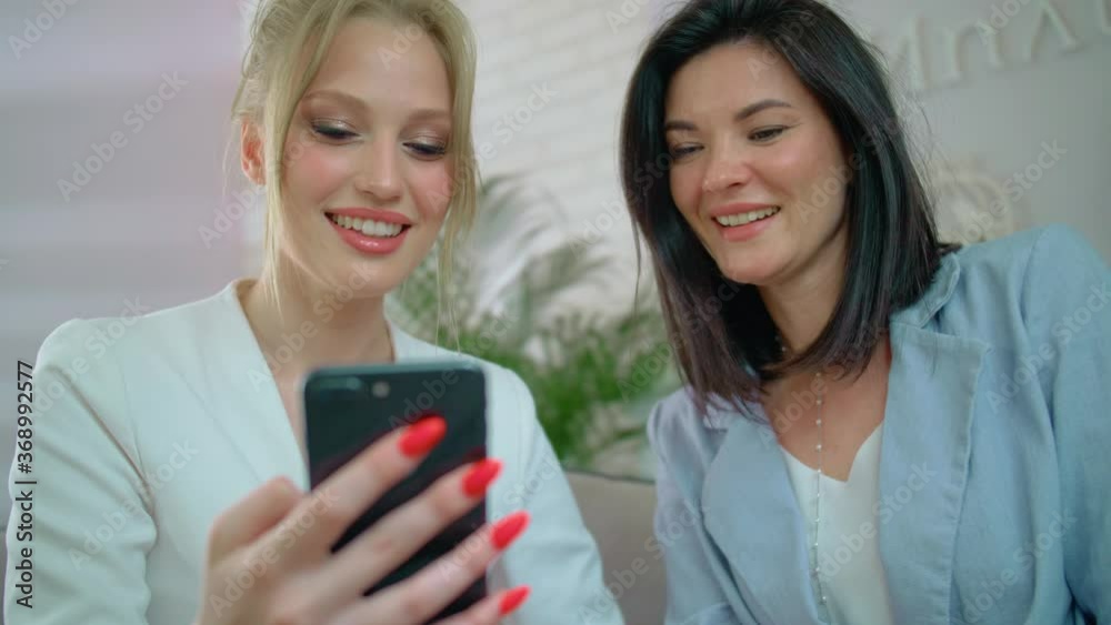 Happy women chat and look at the phone, Girls smile and show each other photos, Friends sit on the couch at home and talk Happy women chat and look at the phone, Girls smile and show each other photos, Friends sit on the couch at home and talk