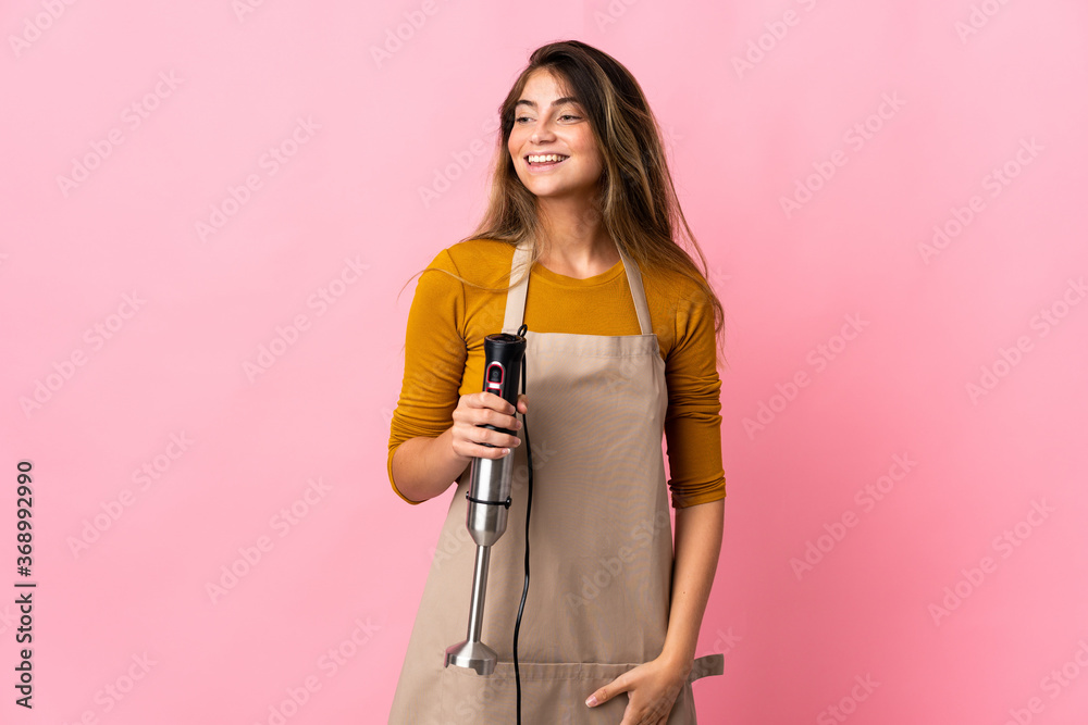 Young chef woman using hand blender isolated on pink background looking to the side and smiling