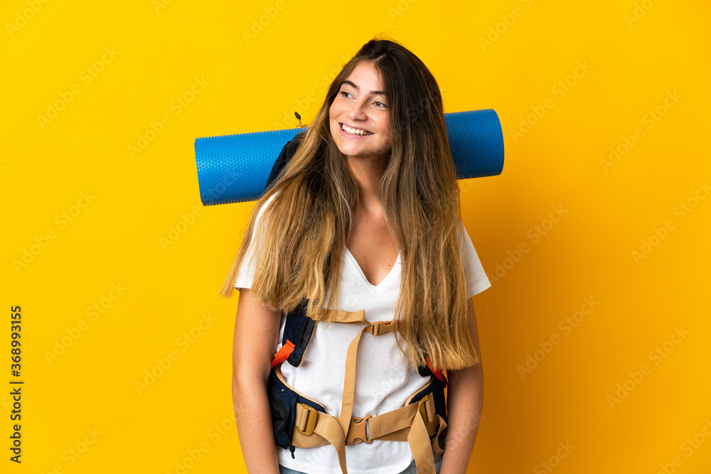 Young mountaineer woman with a big backpack isolated on yellow background looking to the side and smiling Young mountaineer woman with a big backpack isolated on yellow background looking to the side and smiling