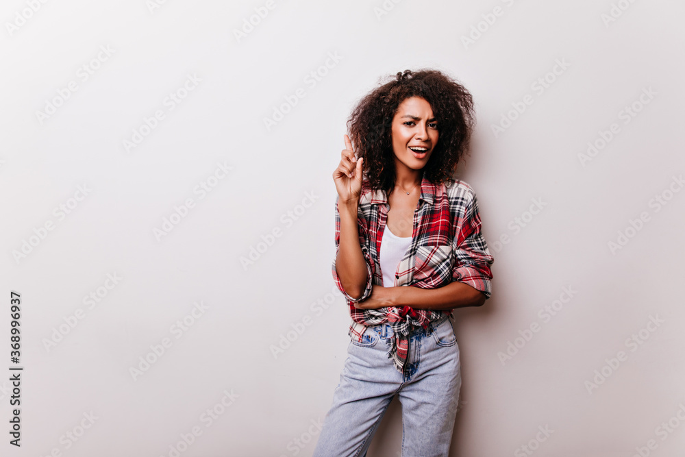 Winsome young lady in blue jeans enjoying indoor photoshoot. Studio portrait of refined african woman in red casual shirt. Winsome young lady in blue jeans enjoying indoor photoshoot. Studio portrait of refined african woman in red casual shirt.