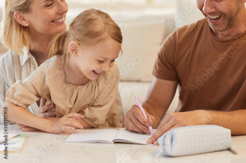 Close up portrait of cute little girl smiling happily with parents helping her draw or study at home, copy space