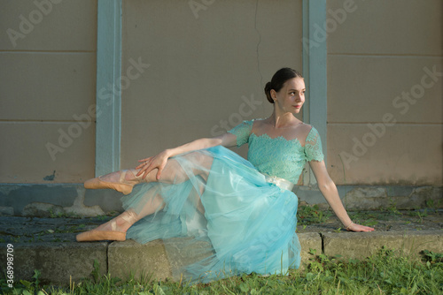 Beautiful graceful young ballerina in pointe shoes and blue dress sitting on the pavement. Female stretching indoors, professional arts. Female fit and healthy lifestyle. Ballet practice on sunset