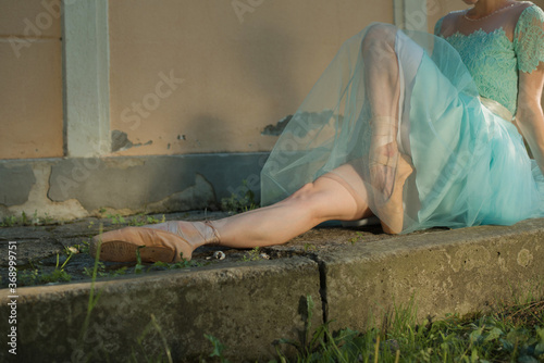 Beautiful graceful young ballerina in pointe shoes and blue dress sitting on the pavement. No face female feet legs stretching indoors toes professional arts. Ballet practice and stretching