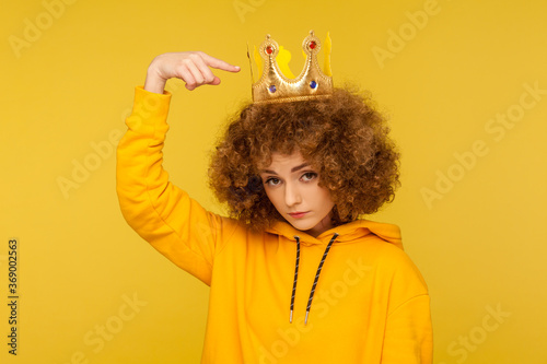 Look, I am best! Selfish haughty curly-haired woman pointing at crown on head and looking with arrogance supercilious, being egoistic with over-inflated ego. studio shot isolated on yellow background