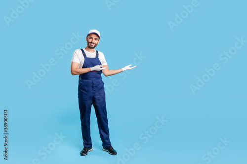 Obraz na plátne Full length cheerful workman in blue overalls, cap and protective gloves standing, showing empty space advertising area