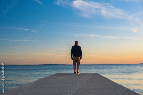 Man in hoodie and short cargo pants standing on dock and looking on sea horizon  island od Pag  Adriatic sea  Croatia 