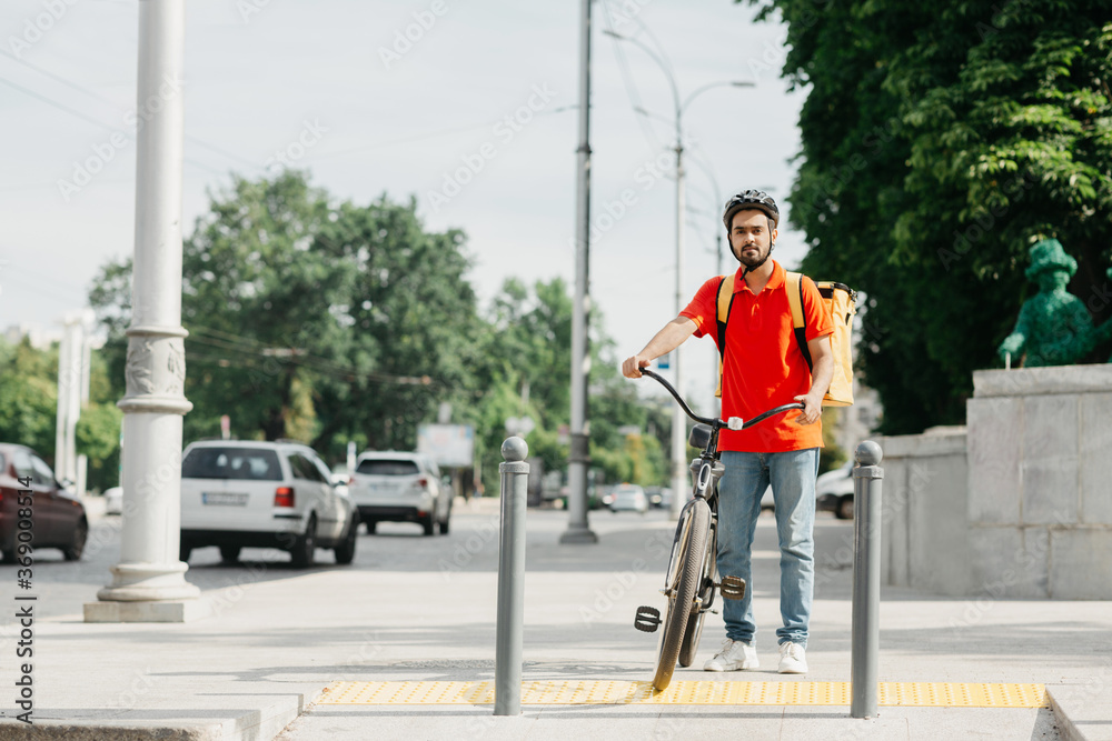 Courier and city traffic. Guy with beard in protective helmet with yellow backpack and bicycle, stands at crossroads