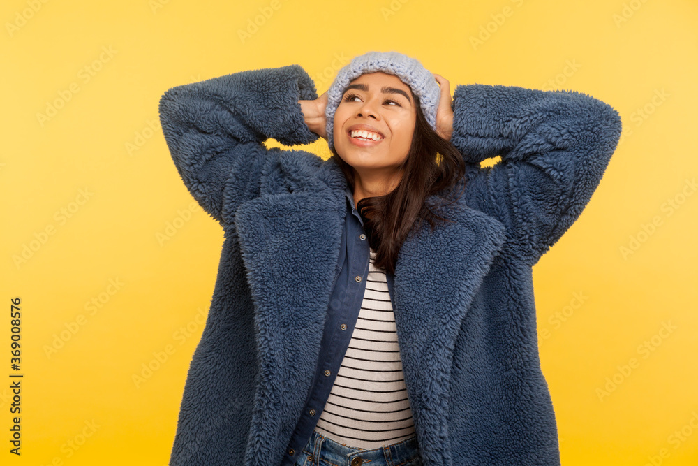 Portrait of carefree happy girl wearing warm winter hat and fur coat looking up smiling joyfully, glamour fashion model in stylish urban outfit enjoying life. studio shot isolated on yellow background