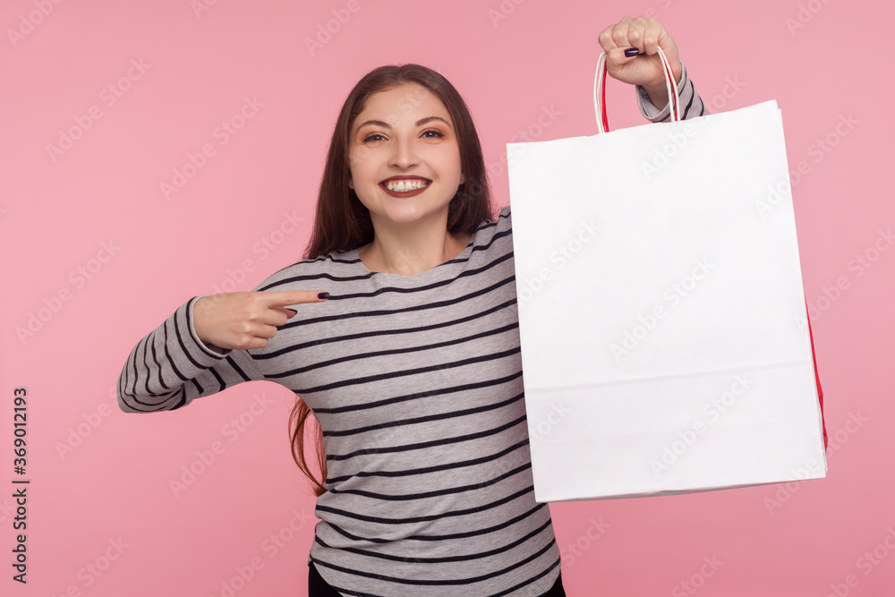 Portrait of joyful lovely woman in striped sweatshirt pointing shopping bag and looking at camera with toothy smile, showing package with blank space for advertise. indoor studio shot, pink background Portrait of joyful lovely woman in striped sweatshirt pointing shopping bag and looking at camera with toothy smile, showing package with blank space for advertise. indoor studio shot, pink background