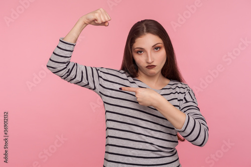 Look how strong I am! Portrait of confident independent woman in striped sweatshirt raising hand pointing biceps, feeling power and energy to achieve goal. studio shot isolated on pink background