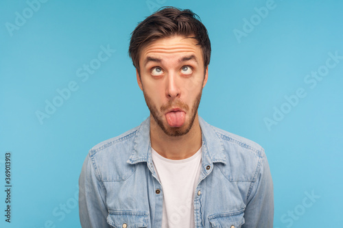Portrait of idiot man in denim shirt making silly humorous face with eyes crossed and tongue out, showing comical silly brainless facial expression. indoor studio shot isolated on blue background © khosrork