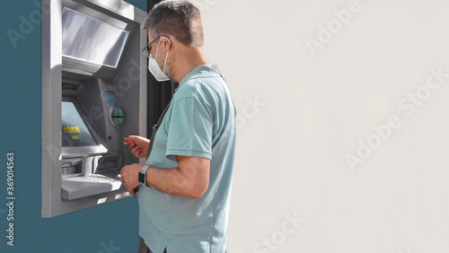A middle aged man with a face mask uses an ATM to withdraw cash. Daily life in epidemic times.