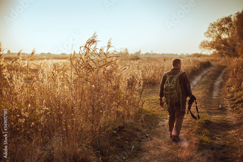 Strong young hunter with red beard holding his gun and walking along the dirt road under blue sky © honey_inside
