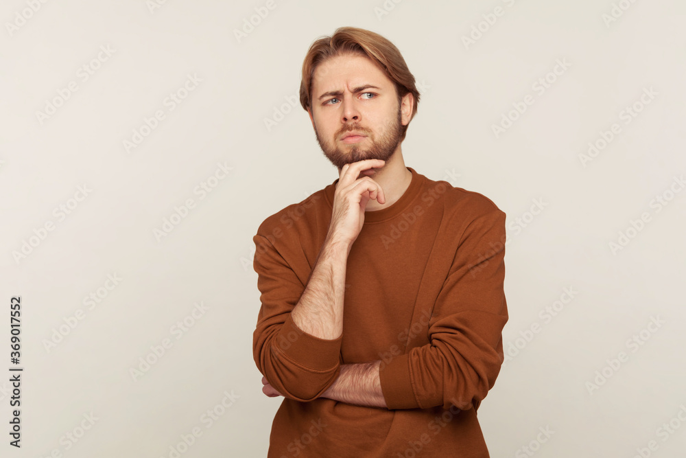 Portrait of thoughtful pensive smart man with neat hair and beard in sweatshirt touching chin while thinking intensely, having doubts, uncertain about choice. studio shot isolated on gray background Portrait of thoughtful pensive smart man with neat hair and beard in sweatshirt touching chin while thinking intensely, having doubts, uncertain about choice. studio shot isolated on gray background