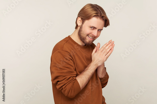 Portrait of sneaky sly scheming man with beard wearing sweatshirt rubbing palm as having cunning evil idea, devious plan in mind, thinking revenge. indoor studio shot isolated on gray background