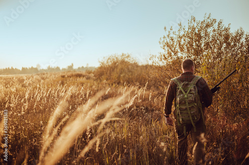 Young hunter in the sunny field with military backpack and shotgun waiting for flying ducks