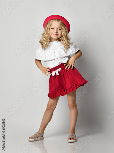 Beautiful positive little blonde girl with curly hair in stylish casual clothing, sandals and hat standing and smiling